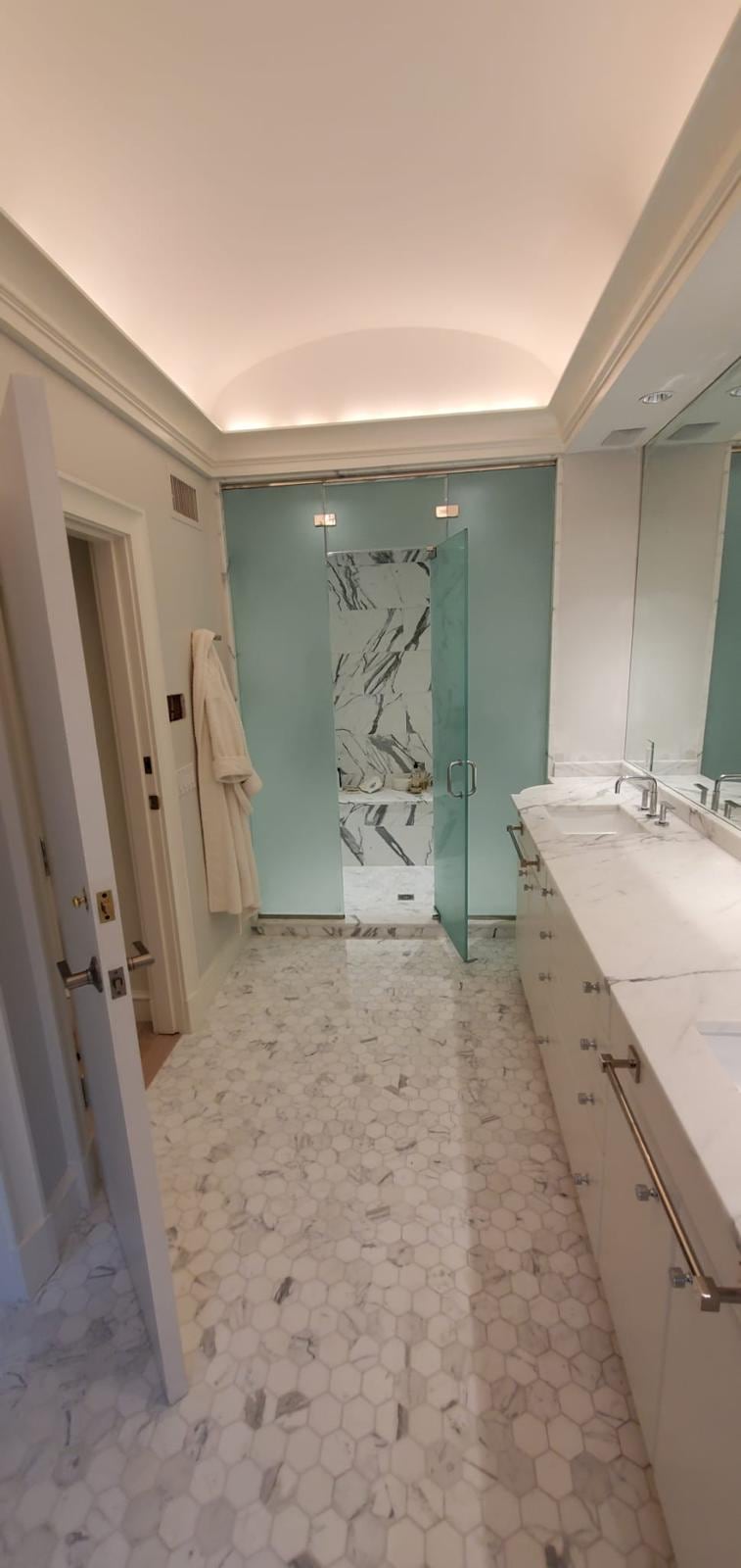 Long bathroom with a double vanity, marble hexagon floor and a frosted glass shower at the end