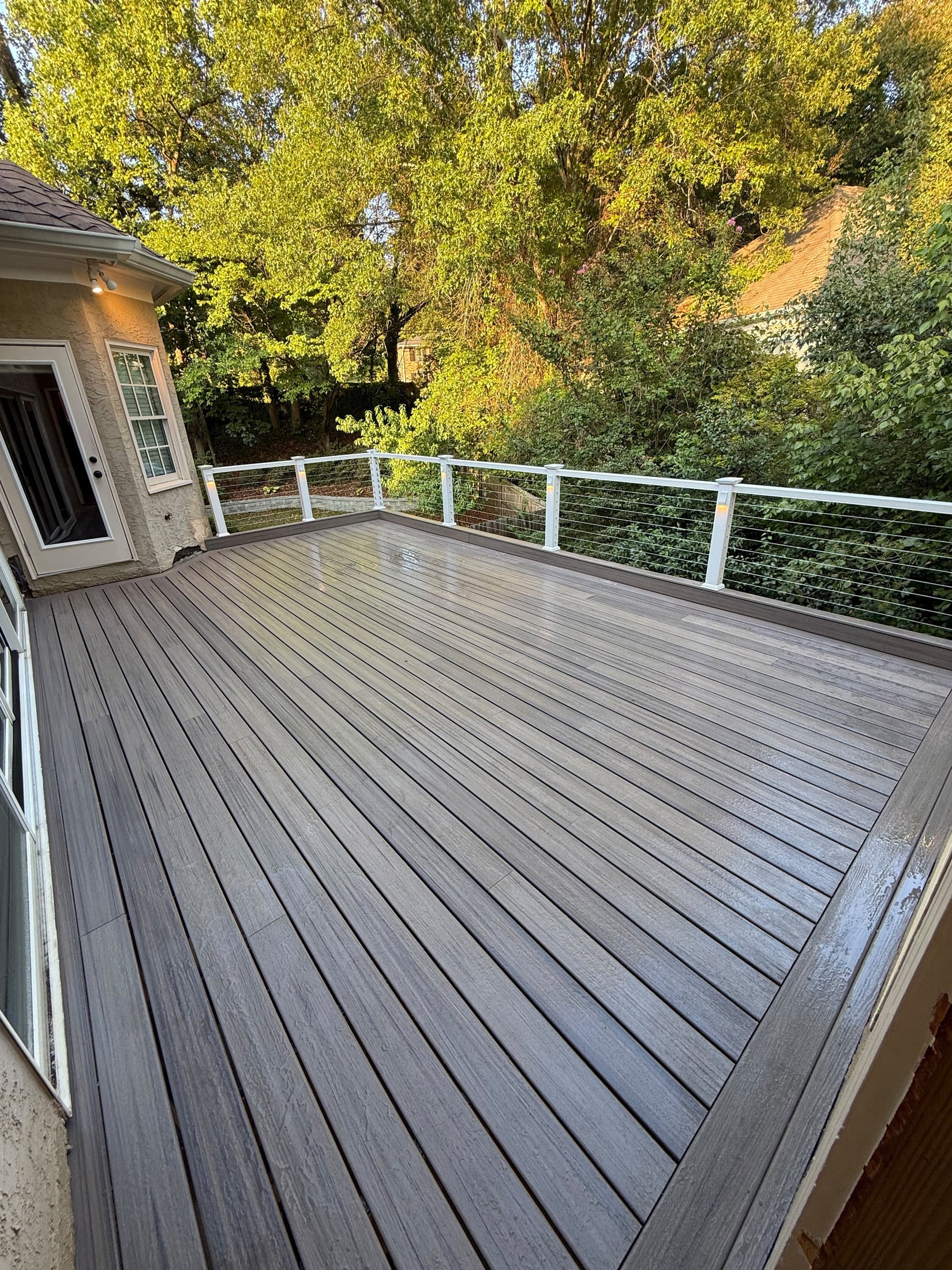 Gray composite deck with white posts and cable railing, seen from the door of the house, trees behind
