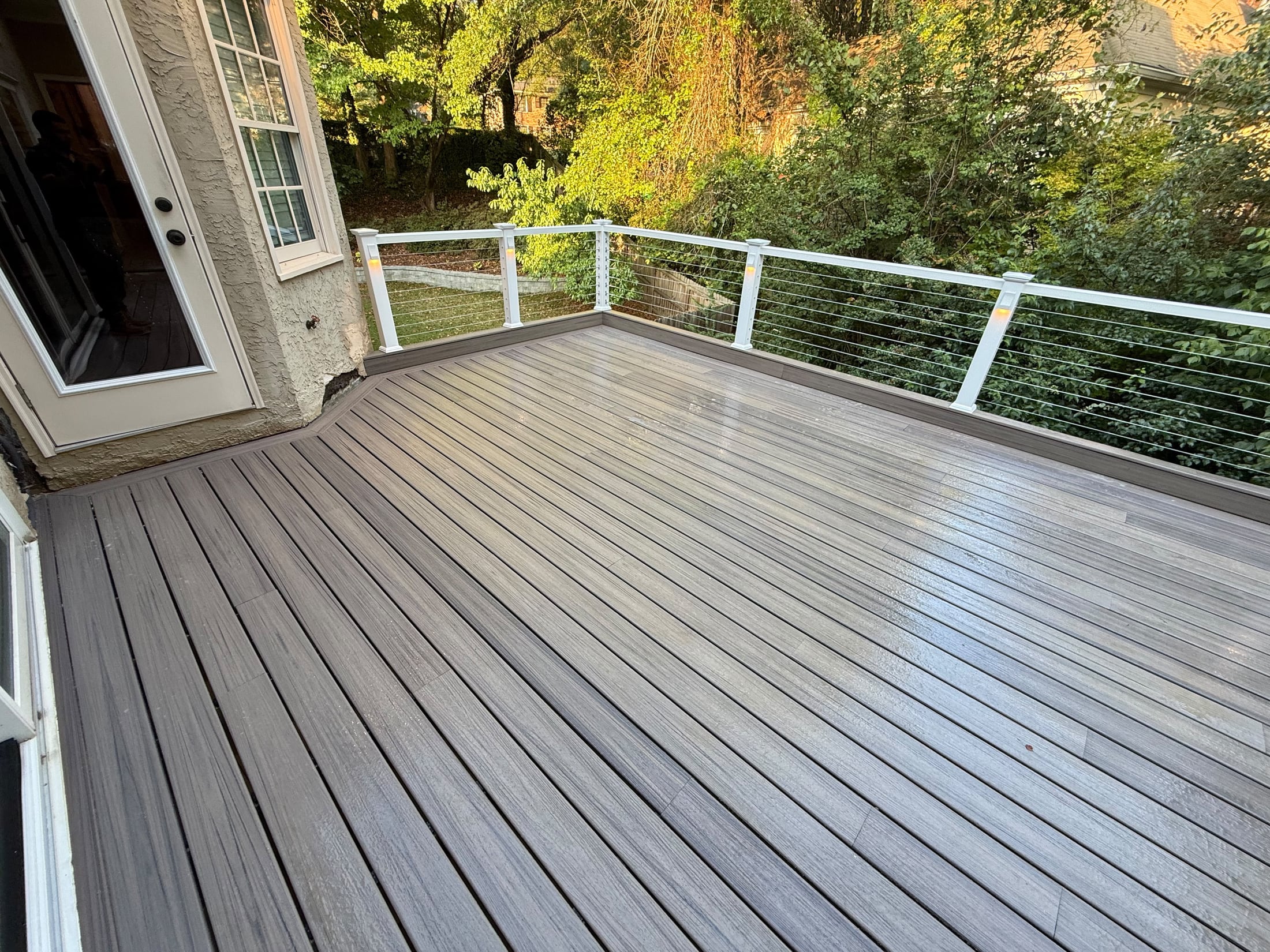 Wide composite deck in gray tones with cable railing along the back edge and woods beyond