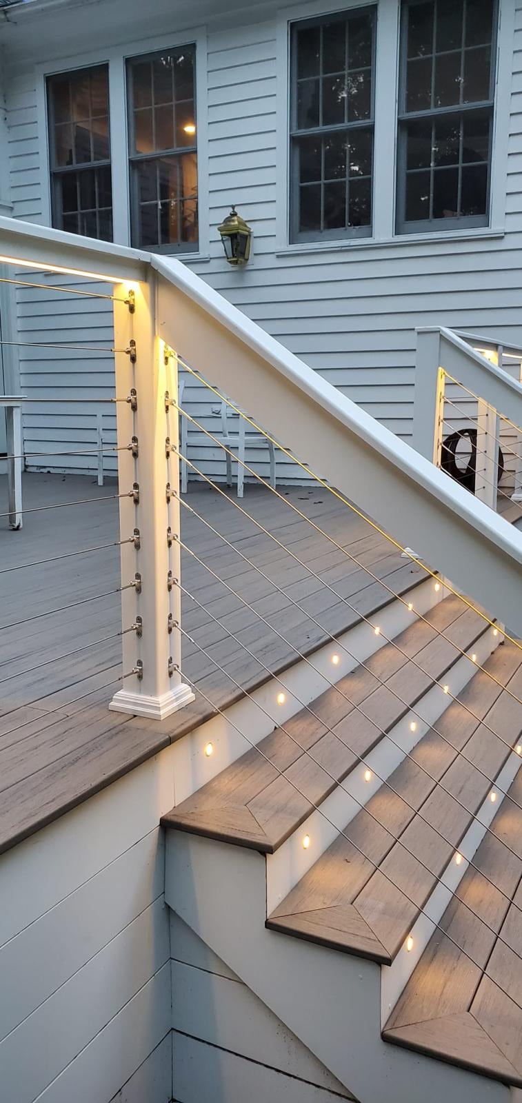 Deck stairs with lit risers and cable railing against a white clapboard house