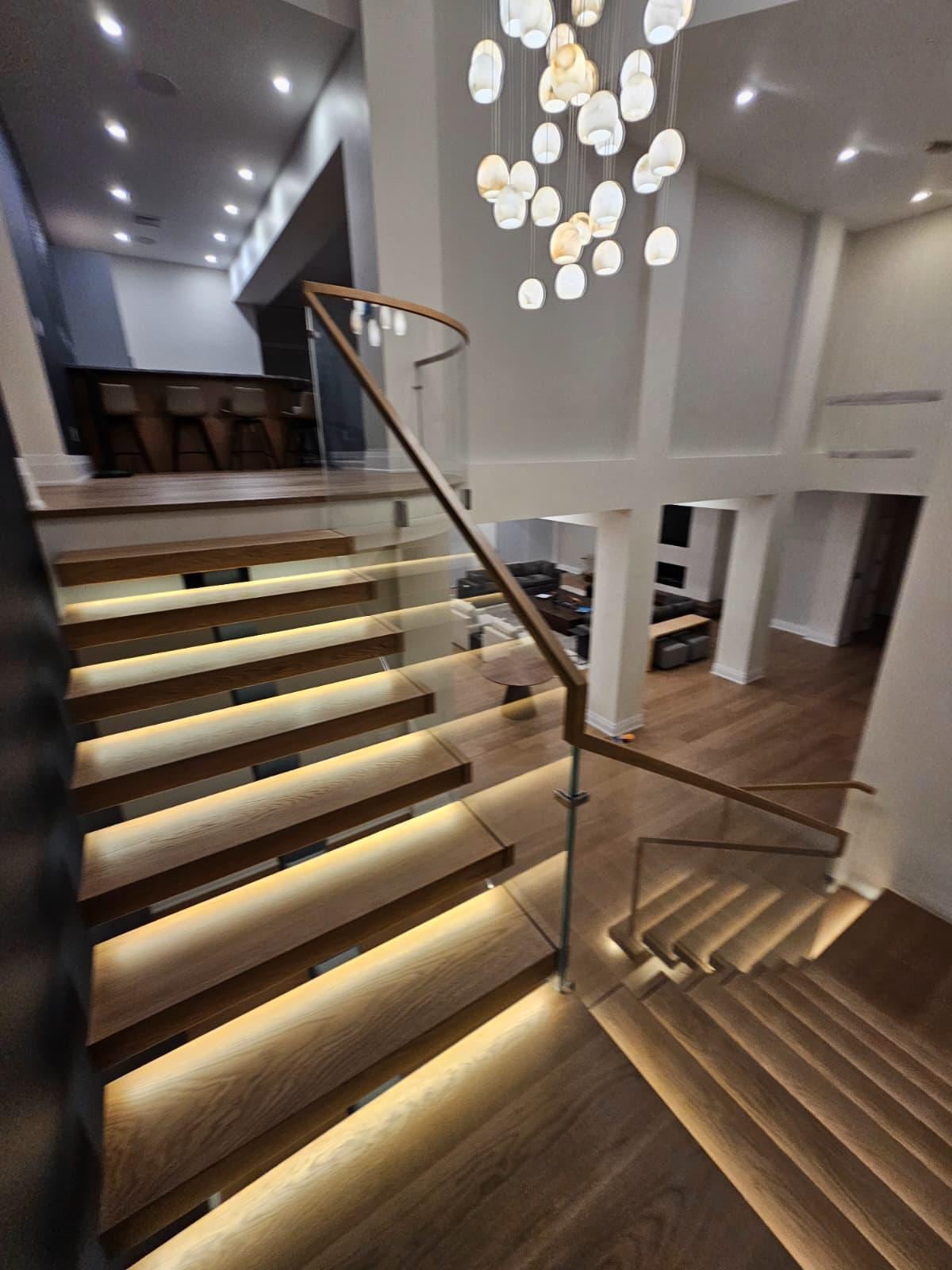 Open-riser wood staircase with under-tread lighting and a glass railing beneath a chandelier