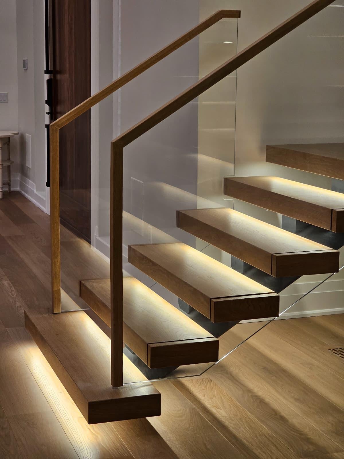 Floating wood stairs with a glass railing and wood handrail, lit from below