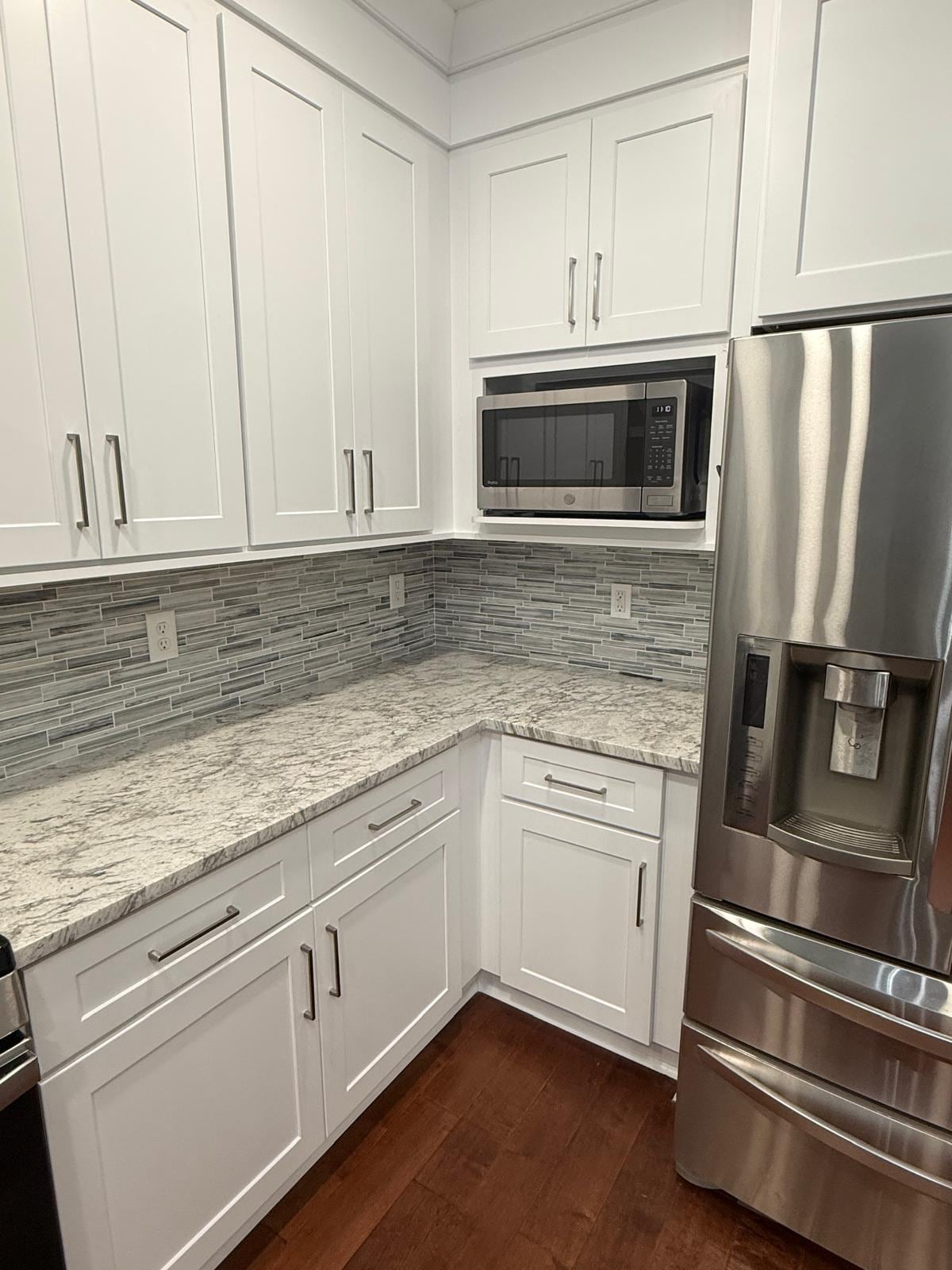 White kitchen cabinets with a built-in microwave and a gray stone backsplash beside a stainless refrigerator