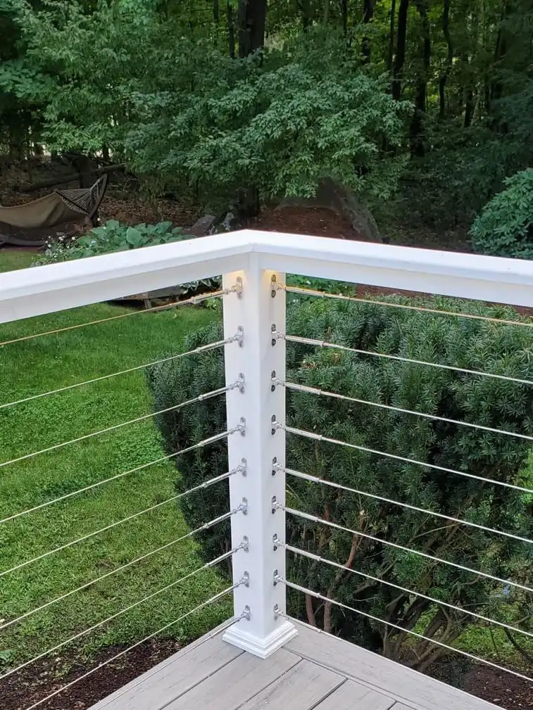 Corner of a gray composite deck with white post and cable railing facing the trees
